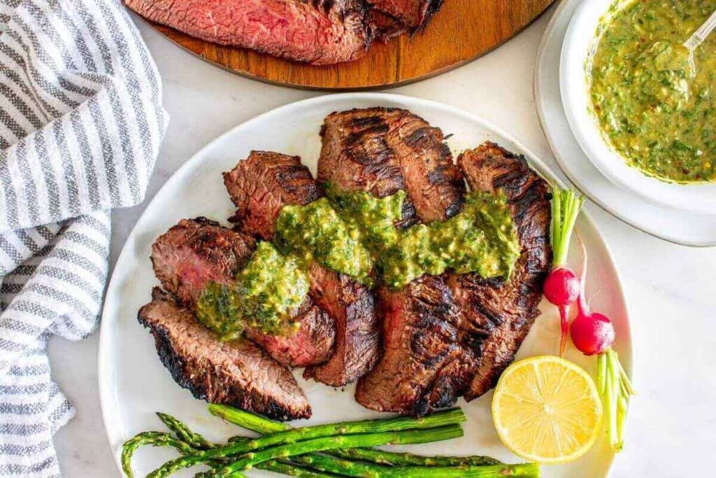 Sliced grilled steak topped with green sauce, served with asparagus, radishes, and a lemon half on a white plate—perfect for a comforting weekend dinner. A bowl of sauce and extra sliced steak are nearby.