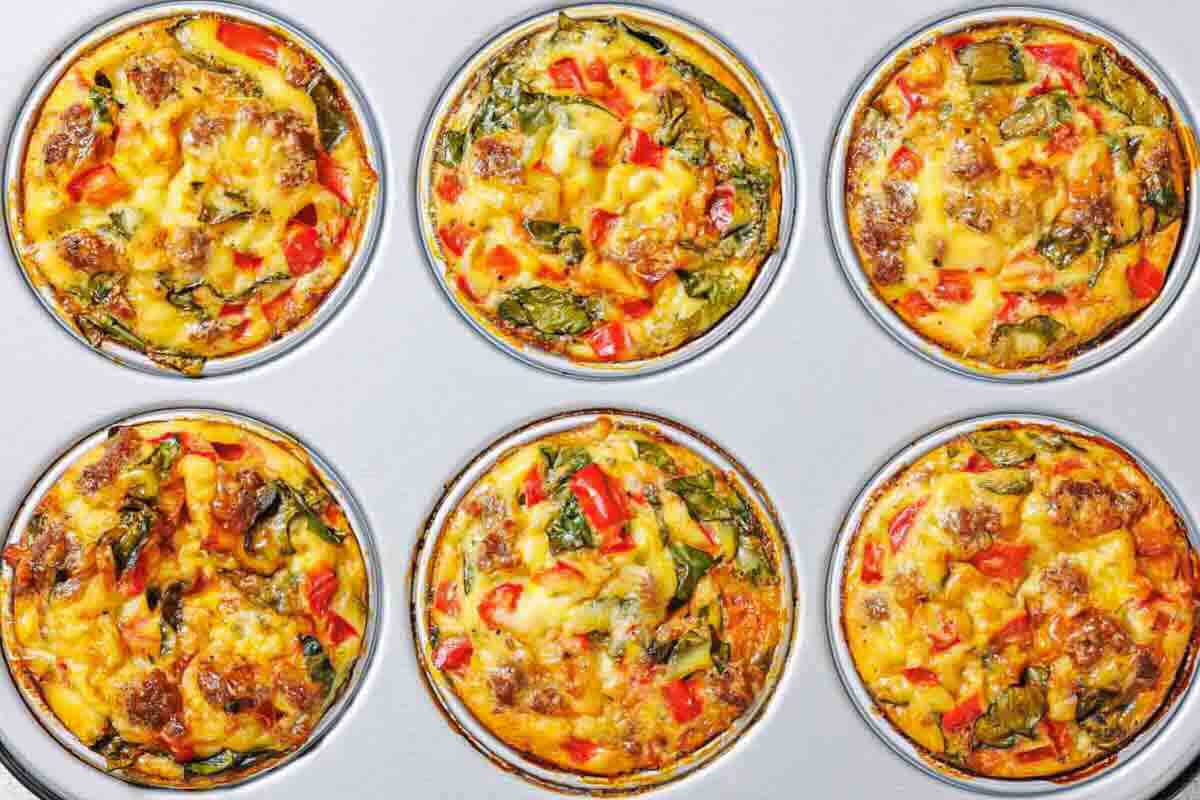 Six baked egg muffins with visible pieces of sausage, spinach, and red bell peppers in a muffin tin.