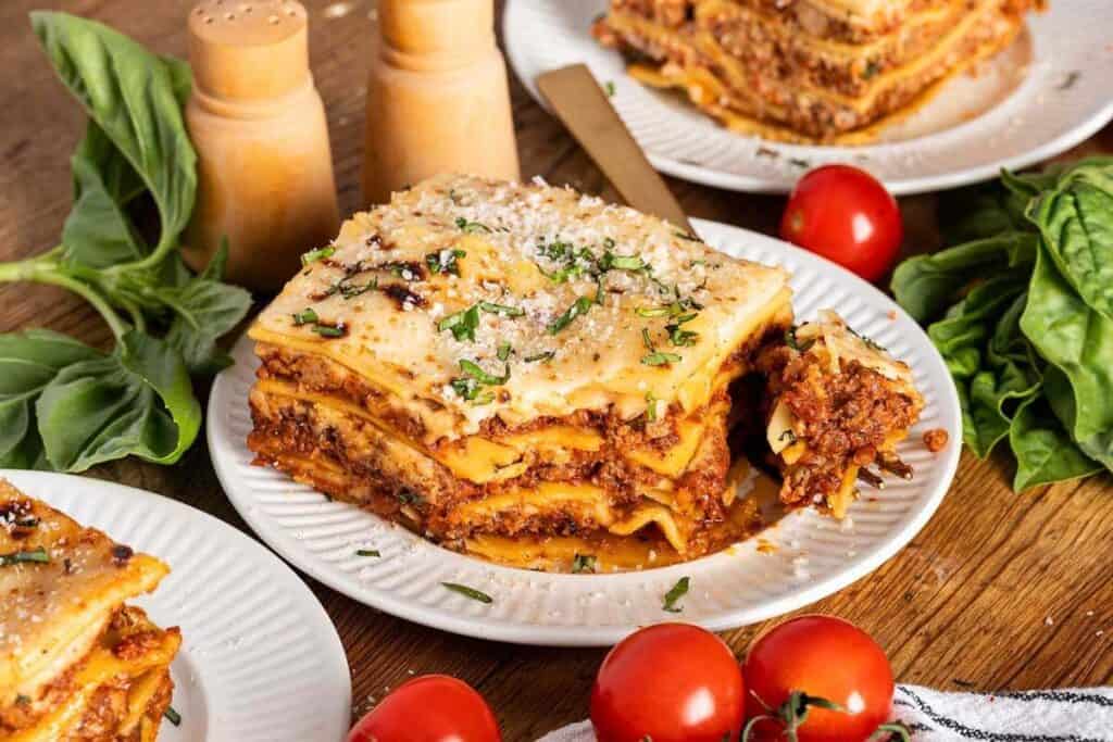A slice of lasagna on a white plate, garnished with herbs and grated cheese, surrounded by fresh basil leaves, tomatoes, and salt and pepper shakers.