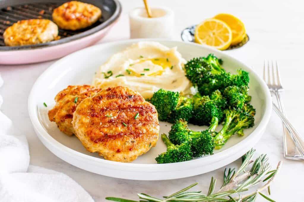 A white plate with two grilled chicken patties, mashed potatoes with butter, and roasted broccoli. A pan with more patties and lemon slices are in the background.