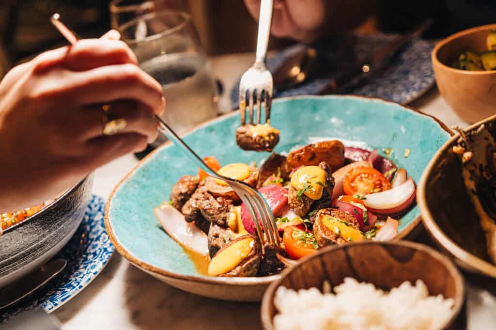 Two people share a meal, using forks to pick from a bowl of roasted vegetables and meat, with a bowl of white rice nearby&mdash;capturing the spirit of Aruba foodie travel 2026.