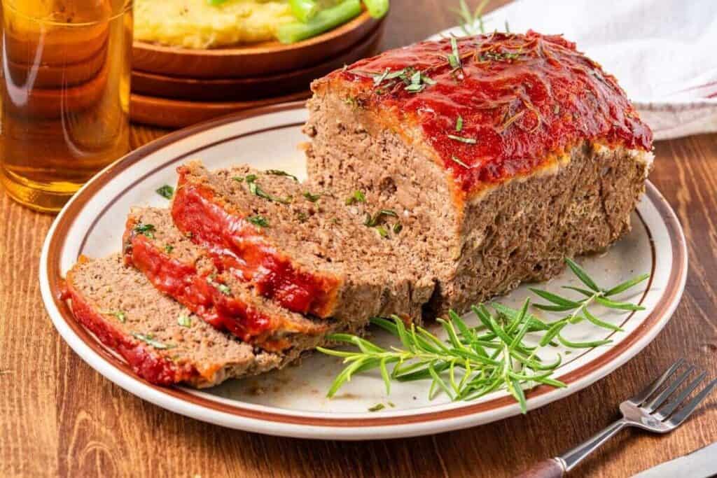 A sliced meatloaf with a tomato glaze is served on a plate and garnished with fresh herbs; a drink and mashed potatoes are in the background.