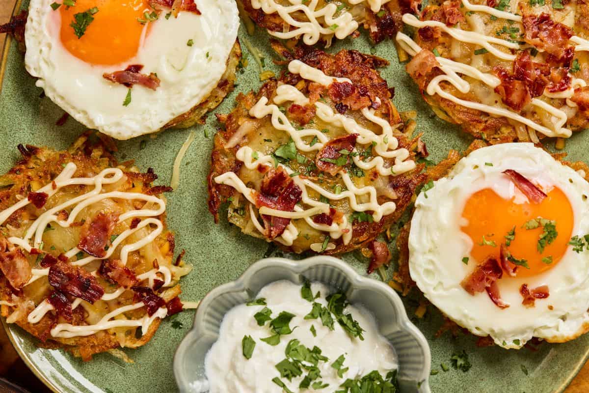 A plate of crispy hash browns topped with fried eggs, chopped bacon, drizzled mayo, and herbs, with a dish of creamy dipping sauce garnished with parsley in the center.