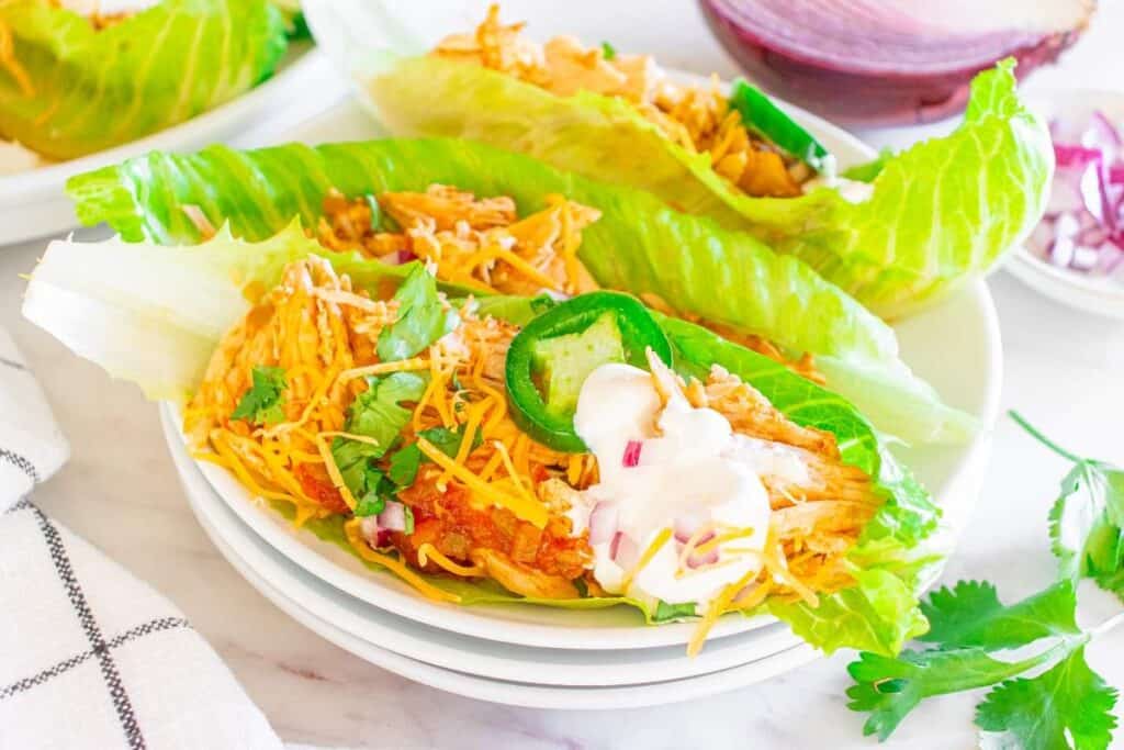 Three lettuce wraps filled with shredded Instant Pot chicken, shredded cheese, sliced jalape&ntilde;o, chopped red onion, cilantro, and a dollop of sour cream on a white plate&mdash;perfect for fans of quick instant pot dishes.