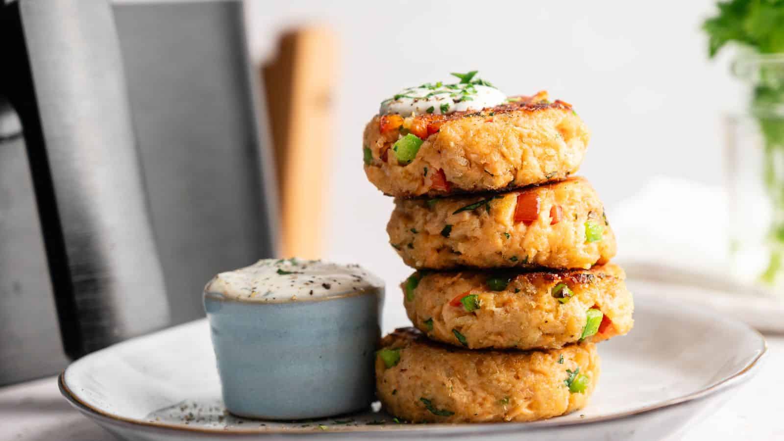 A stack of four vegetable patties garnished with herbs is arranged on a plate next to a small cup of creamy dipping sauce.