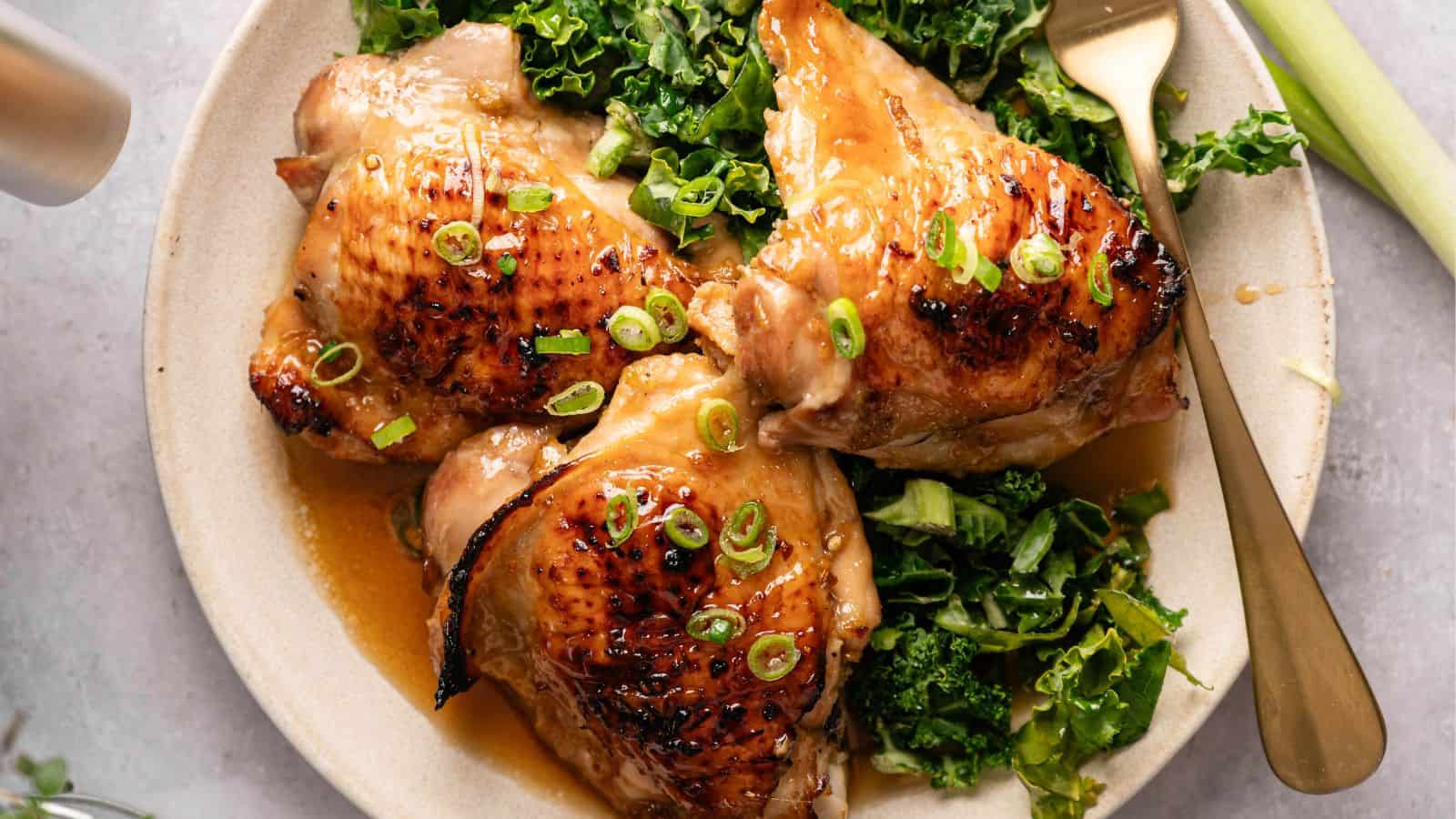Three glazed chicken thighs garnished with chopped green onions, served on a bed of leafy greens on a round plate with a fork.