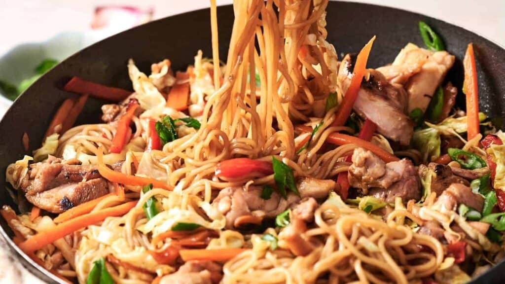 A pan of stir-fried noodles with sliced chicken, carrots, bell peppers, cabbage, and green onions being lifted with chopsticks.