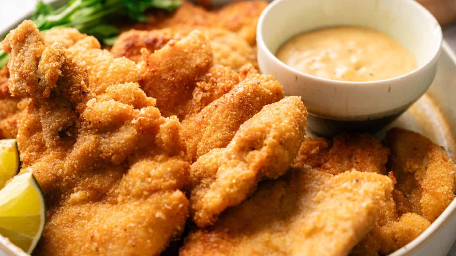 A plate of breaded and fried chicken cutlets served with lemon wedges, a bowl of dipping sauce, and greens in the background.