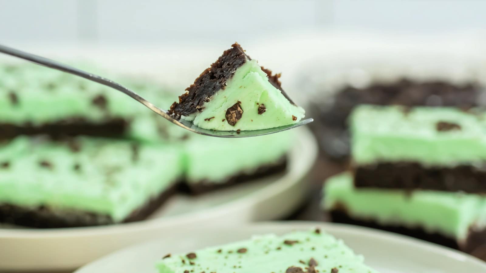 A fork holds a square piece of mint chocolate dessert with a green layer and chocolate crust, with more pieces on plates in the background.