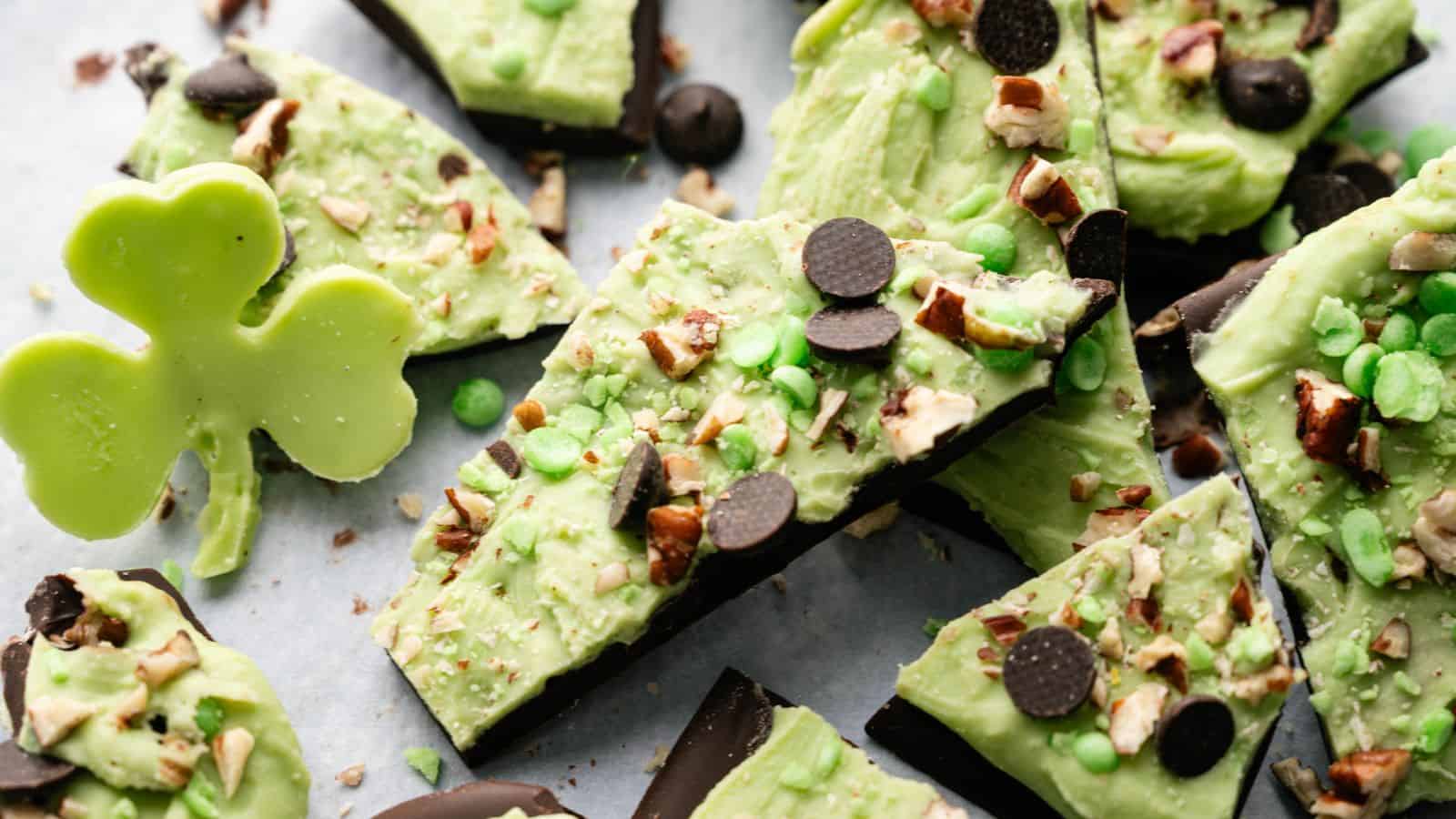 Pieces of green and dark chocolate bark topped with nuts and chocolate chips are scattered on a surface. A shamrock-shaped green chocolate sits on the left side.