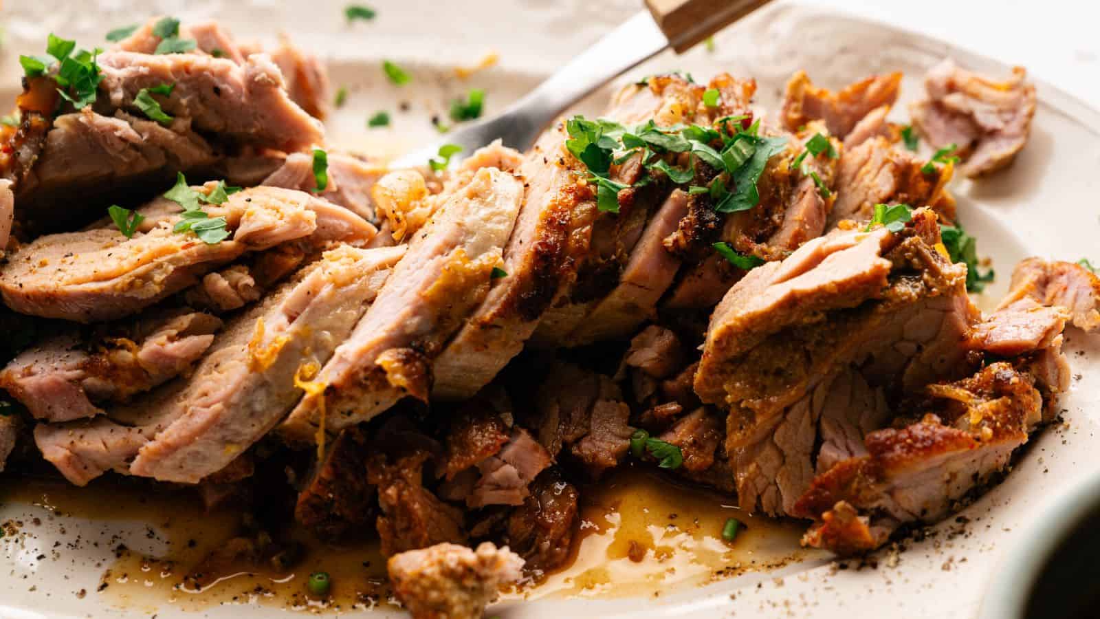 Sliced roasted pork garnished with chopped herbs is arranged on a plate, with visible juices and a serving fork nearby.
