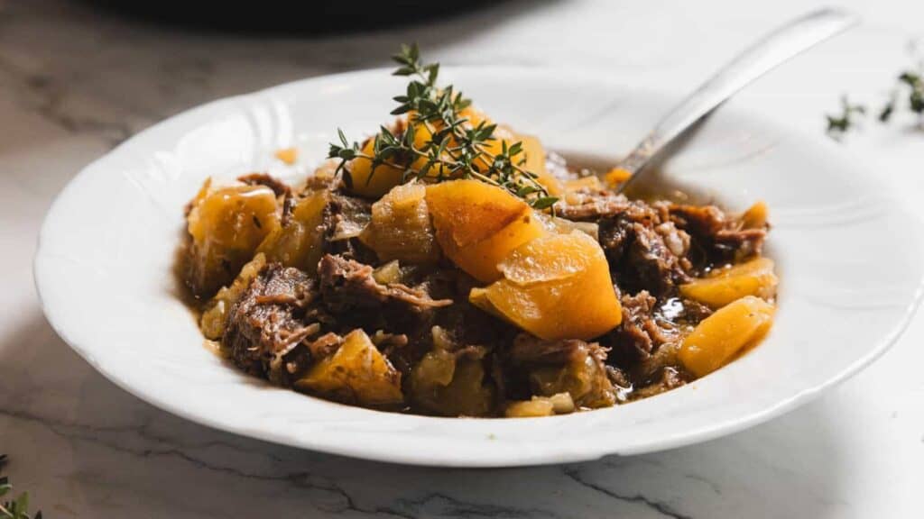 A white bowl filled with beef stew featuring chunks of meat and potatoes, garnished with a sprig of fresh thyme, with a spoon resting in the dish.