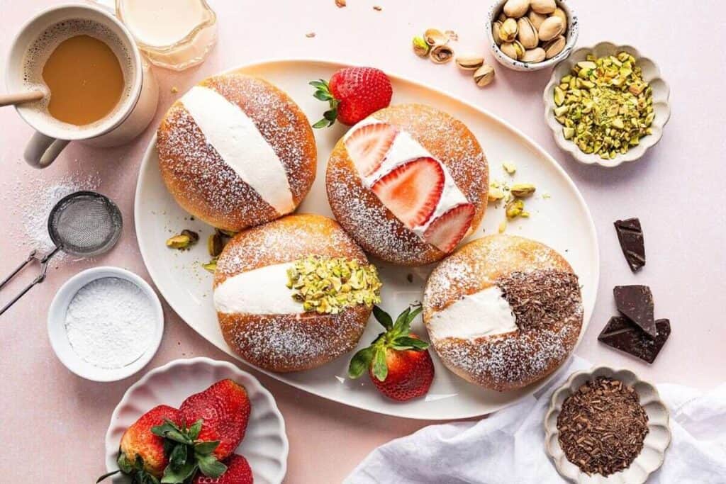 A plate of four filled doughnuts topped with powdered sugar, cream, strawberries, pistachios, and chocolate—an irresistible treat for those who love sweet breakfast recipes—surrounded by fresh ingredients and a cup of coffee.