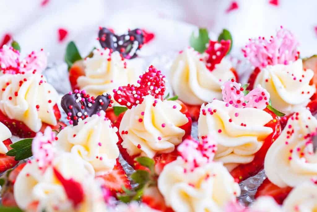 Close-up of Valetines Treats topped with swirls of white frosting, red and pink candy decorations, and colorful sprinkles, beautifully arranged on a tray.