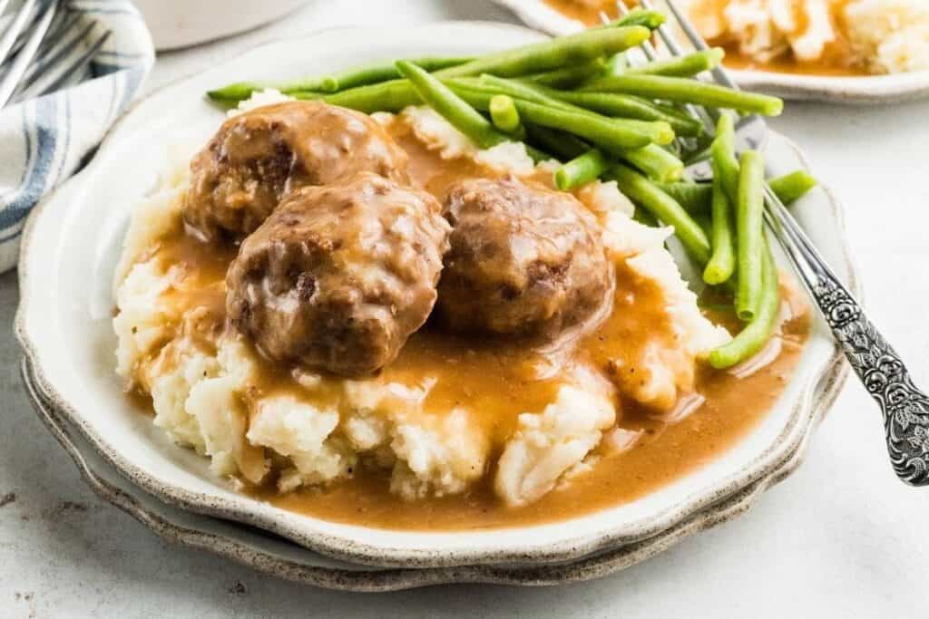 A plate with mashed potatoes, three meatballs topped with brown gravy, and a side of green beans&mdash;one of the tastiest beef recipes you&rsquo;ll want to try.