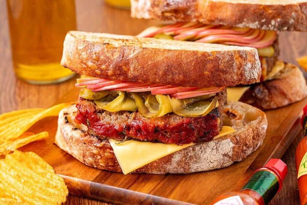 A perfect workday meal idea: a sandwich with a meat patty, sliced pickles, cheddar cheese, and apple slices on rustic bread, served with potato chips and bottles of hot sauce.