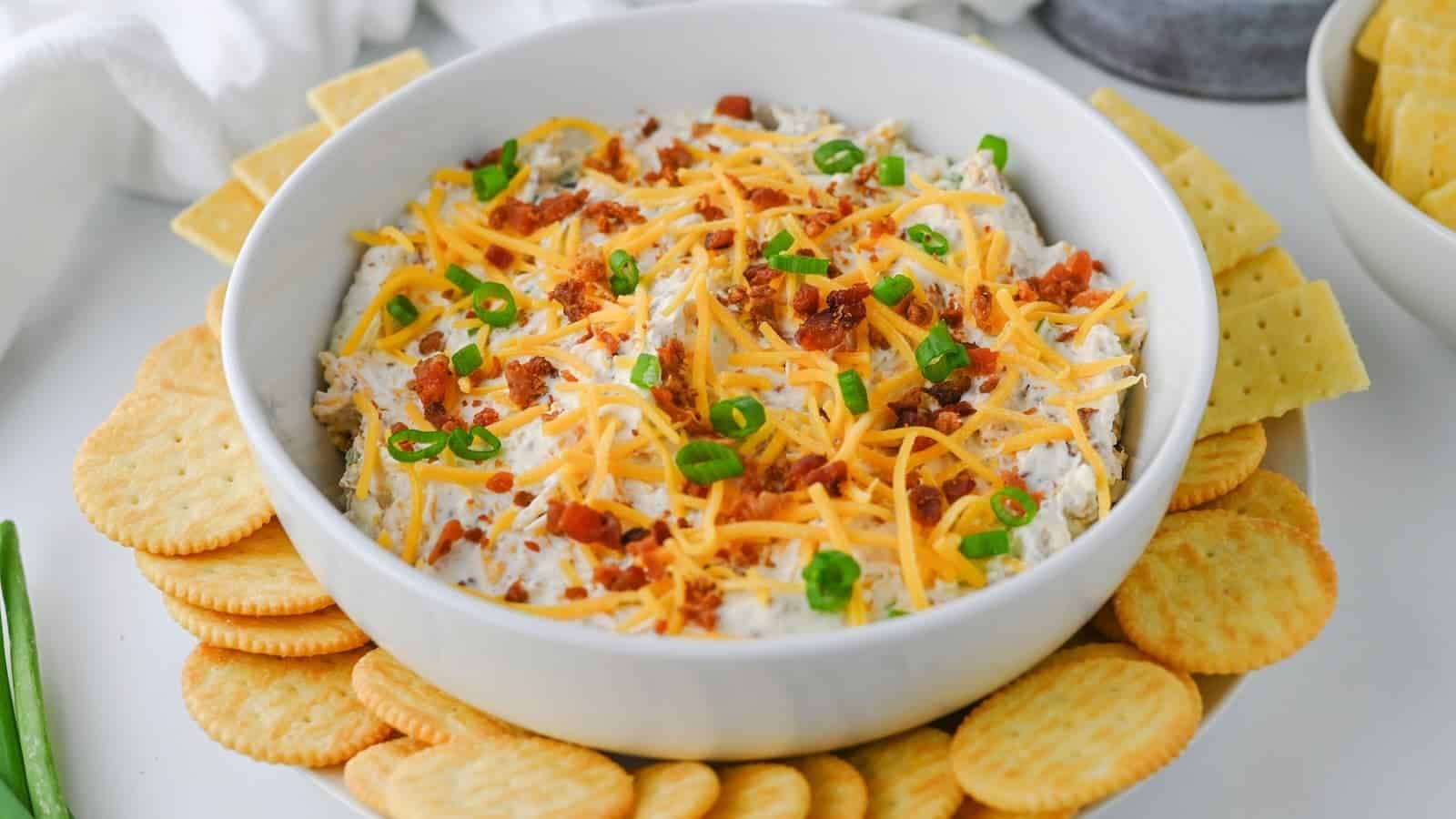 A bowl of creamy million dollar cheese dip topped with shredded cheese, bacon bits, and chopped green onions, surrounded by round crackers.