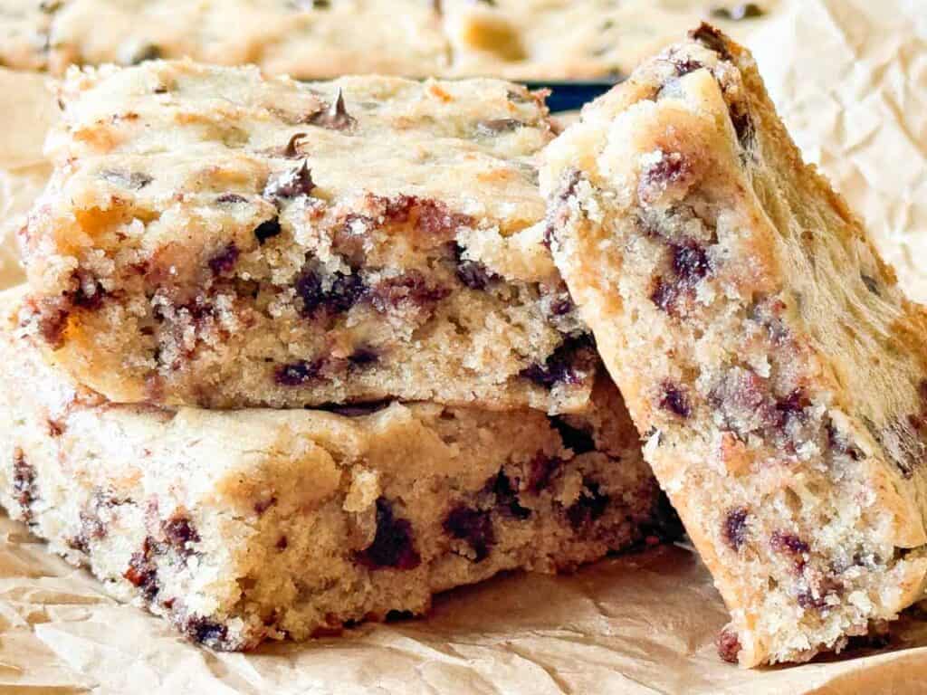 Three thick chocolate chip cookie bars are stacked on brown parchment paper, showing their soft, crumbly texture and visible chocolate chips throughout.