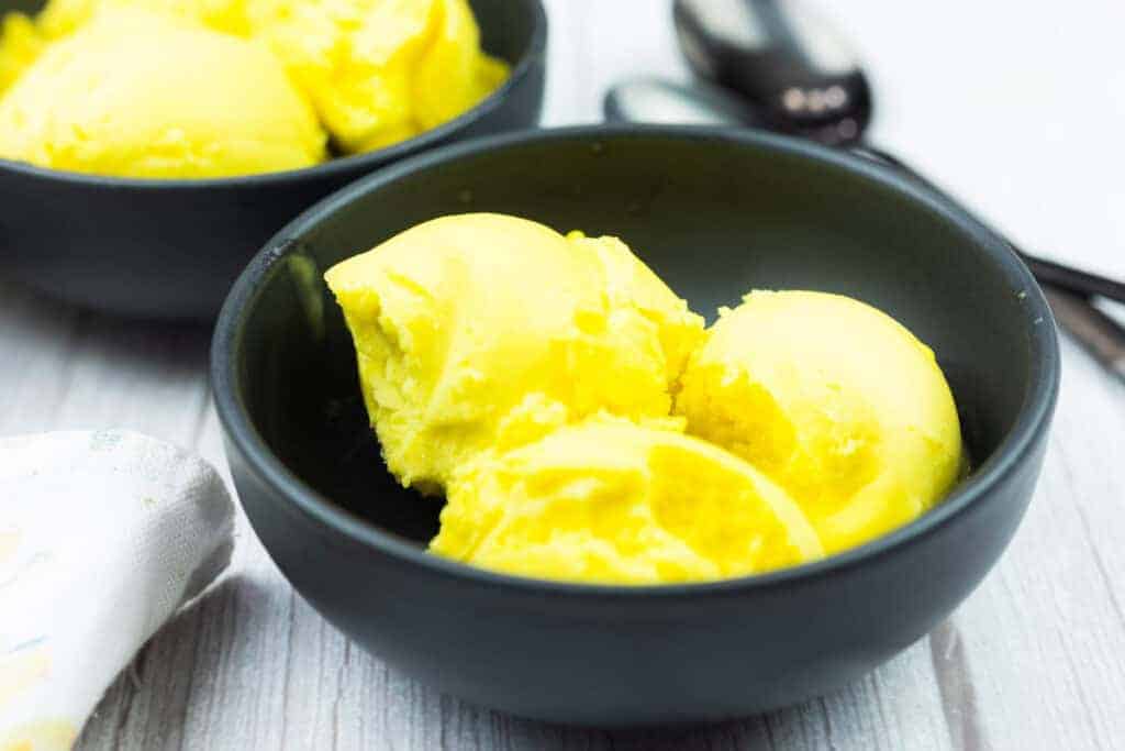 A black bowl containing three scoops of bright yellow ice cream sits on a light wooden surface, with another bowl and metal spoons in the background.