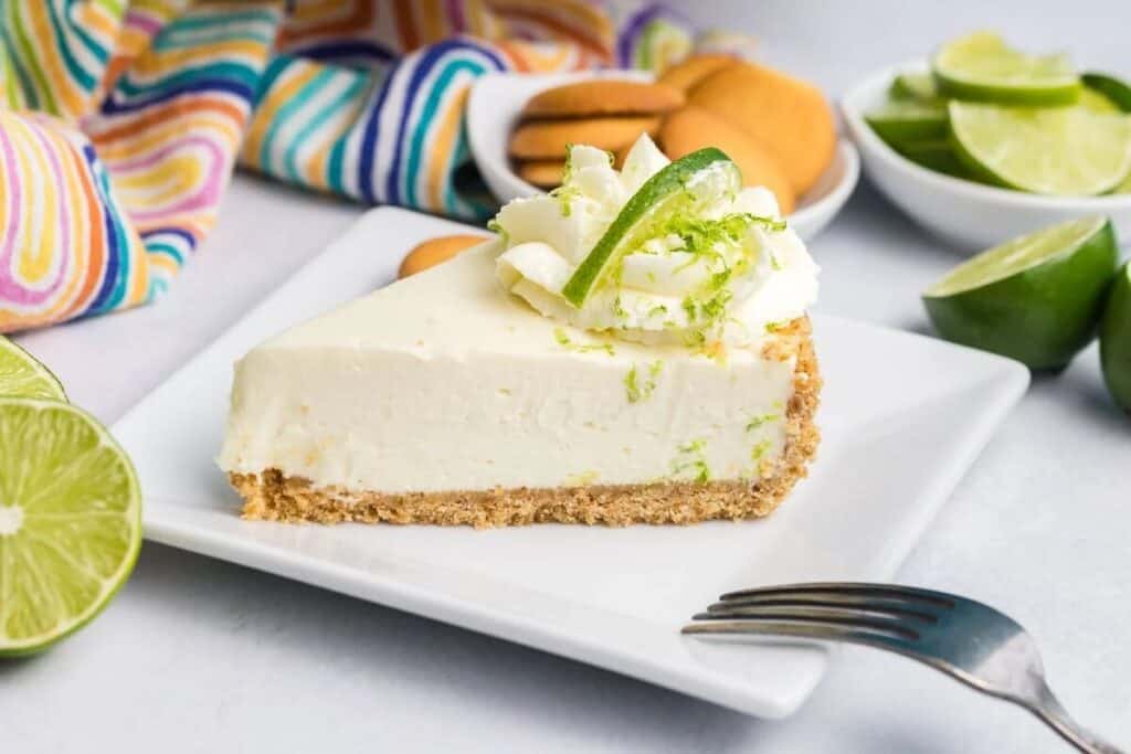 A slice of key lime pie with whipped cream and lime zest on top, served on a square white plate with a fork, lime halves, and cookies in the background&mdash;perfect for fans of simple cheesecake desserts.