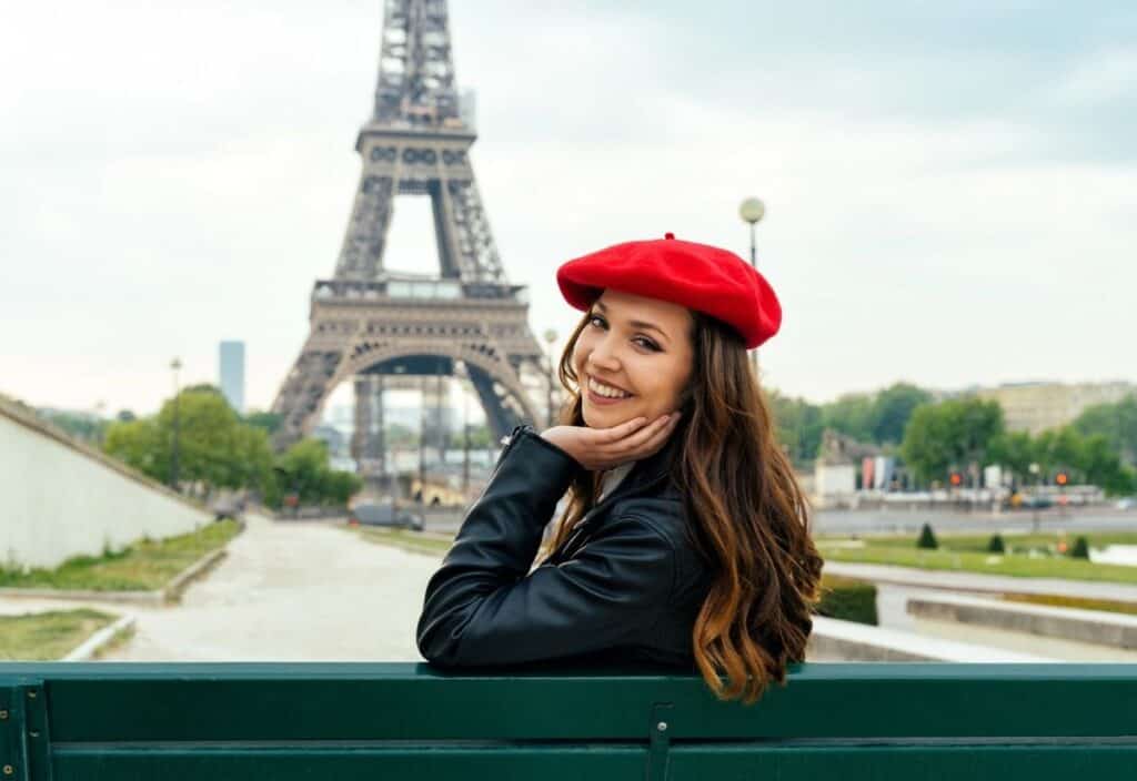 A woman wearing a red beret and black jacket sits on a bench, smiling, with the Eiffel Tower visible in the background, capturing that chic Emily in Paris lifestyle.