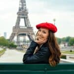 A woman wearing a red beret and black jacket sits on a bench, smiling, with the Eiffel Tower visible in the background, capturing that chic Emily in Paris lifestyle.