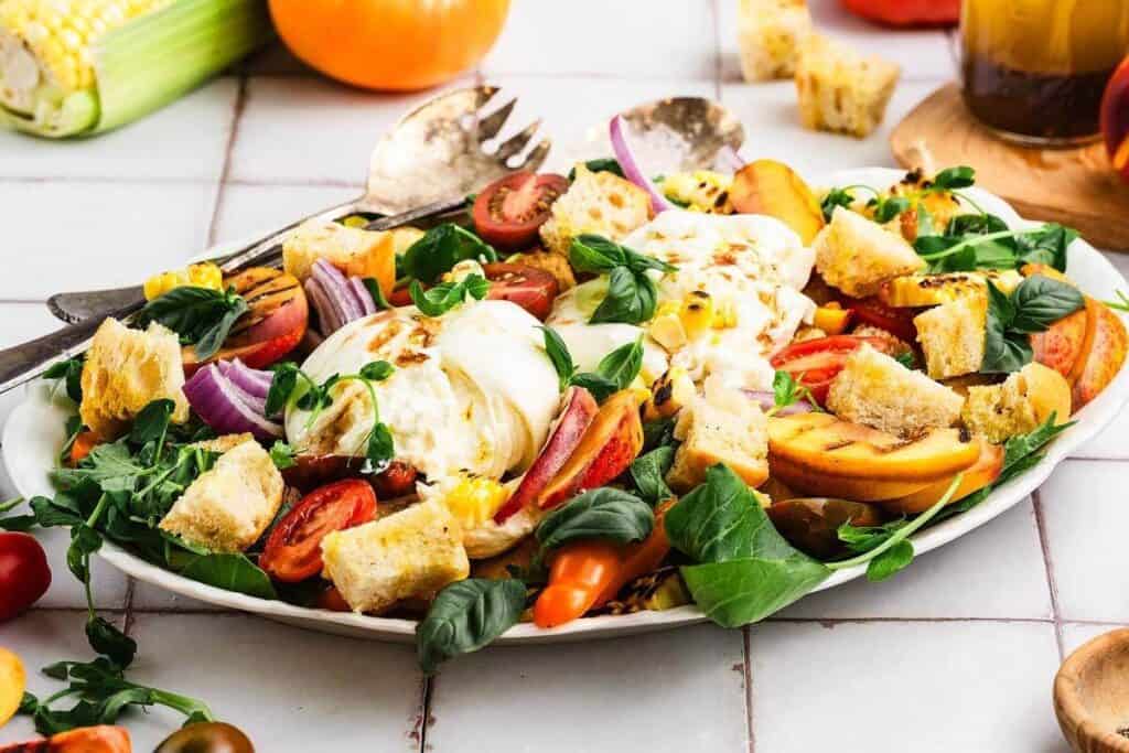 A white platter holds a salad with tomatoes, grilled peaches, red onion, fresh greens, mozzarella, corn, and croutons&mdash;an inspiring option for workday meal ideas&mdash;served with utensils on the side.