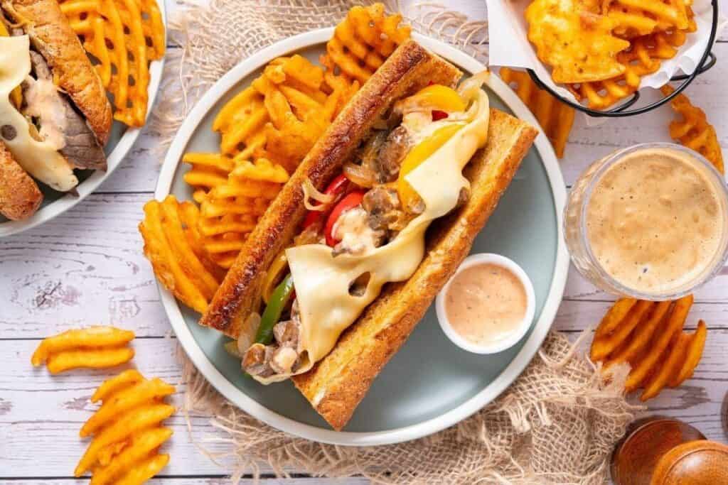 A plate with a cheesesteak sandwich&mdash;one of the tastiest beef recipes&mdash;topped with melted cheese, waffle fries, dipping sauce, and a glass of light beer sits on a wooden table.