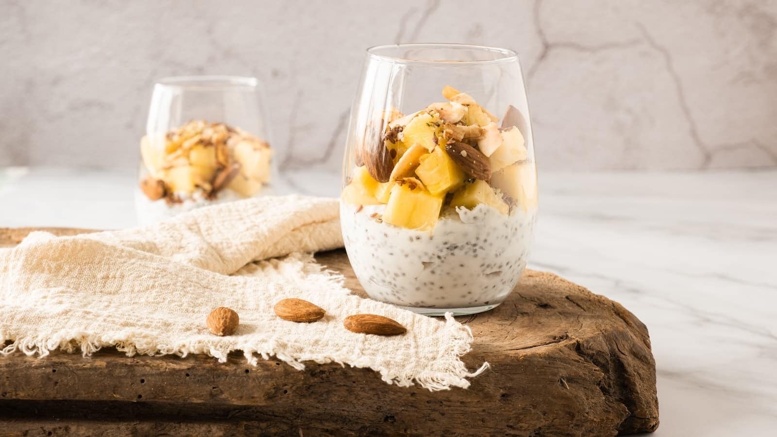 Two glasses filled with chia pudding, topped with diced pineapple, coconut flakes, and nuts, placed on a wooden board with a textured cloth and a few almonds.