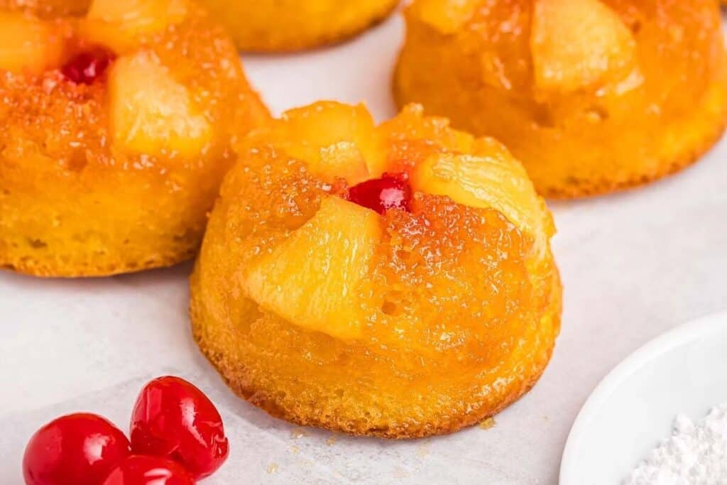 Mini pineapple upside-down cakes topped with pineapple chunks and a cherry, placed on a light surface with extra cherries nearby.