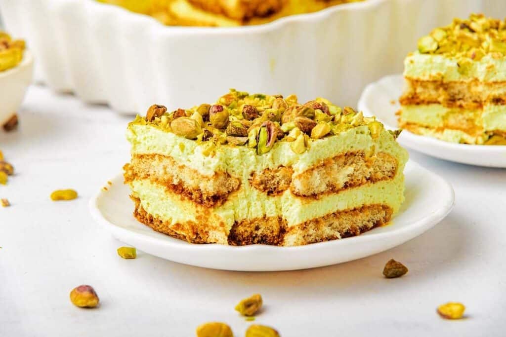 A slice of layered pistachio tiramisu, a delightful no bake treat, is topped with chopped pistachios on a white plate, with more pistachios scattered around.