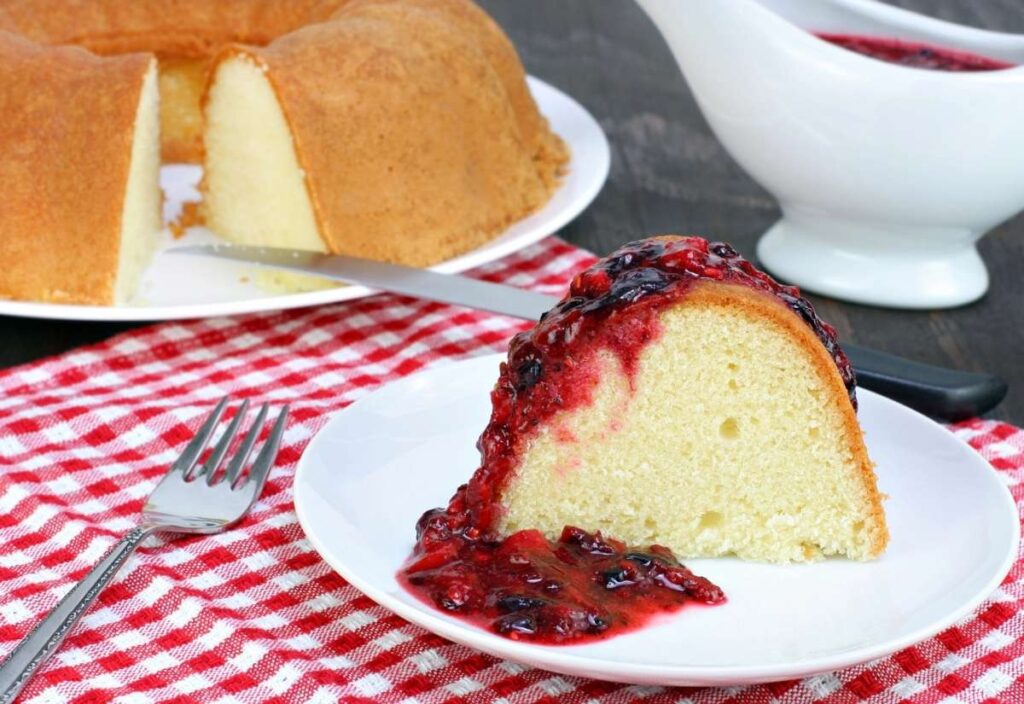 A slice of pound cake with berry sauce on a white plate, next to a fork. The rest of the cake and sauce are in the background on a red checkered cloth.