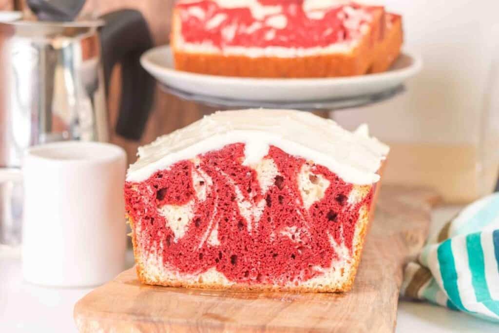 A sliced marble loaf cake with red and white swirls, topped with white icing, sits on a wooden board; another slice is on a plate in the background&mdash;perfect for fans of Starbucks Copy Cat Recipes.