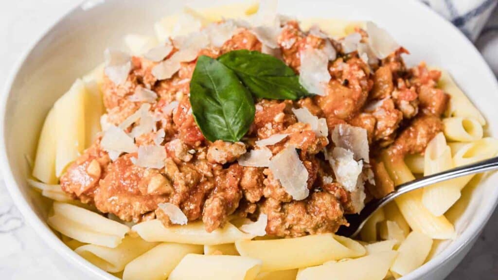 A bowl of penne pasta topped with meat sauce, grated cheese, and a basil leaf, with a spoon resting on the side.