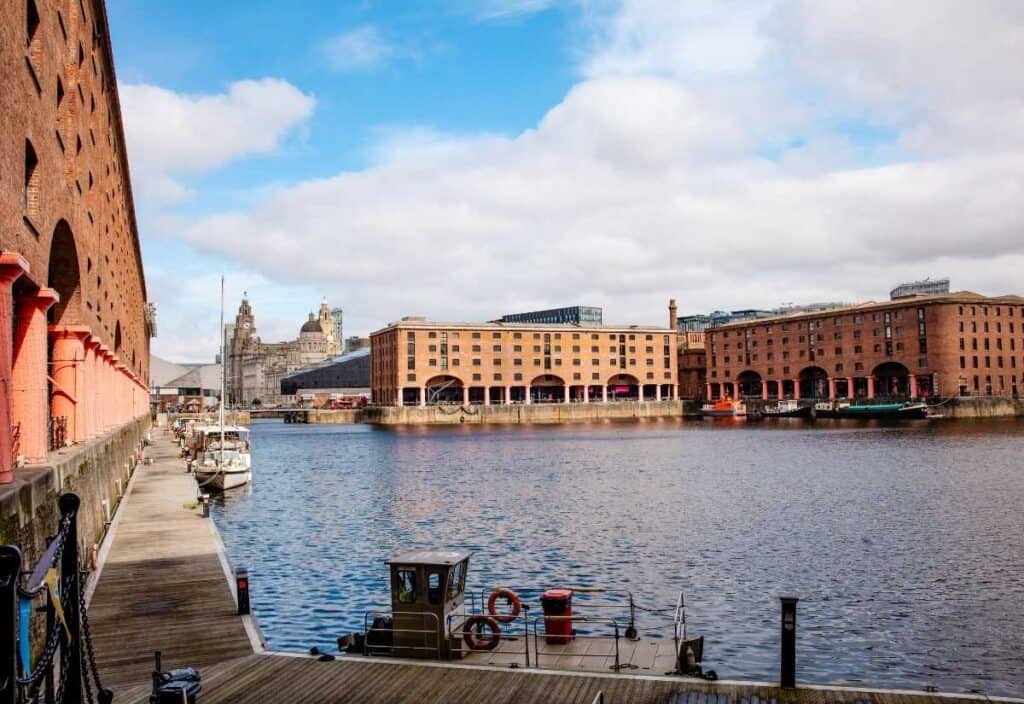 A waterfront scene along the Liverpool waterfront, showing historic red-brick buildings surrounding a dock with boats, under a partly cloudy sky.