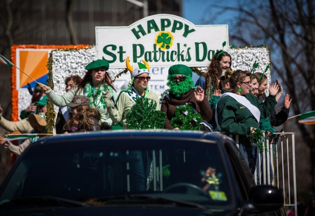 People ride a decorated float with "Happy St. Patrick's Day" sign, wearing green outfits and accessories, during a parade. A black vehicle is visible in the foreground.