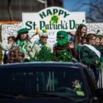 People ride a decorated float with "Happy St. Patrick's Day" sign, wearing green outfits and accessories, during a parade. A black vehicle is visible in the foreground.