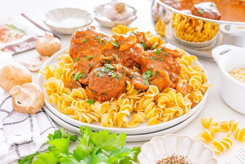 A plate of egg noodles topped with meatballs in tomato sauce, garnished with herbs. Surrounding the plate are mushrooms, parsley, a bowl of pepper, and a pot of sauce.