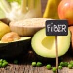 Assorted high-fiber foods including avocado, apple, whole grain bread, green peas, and grains, with a small chalkboard sign labeled “FIBER” in the center.