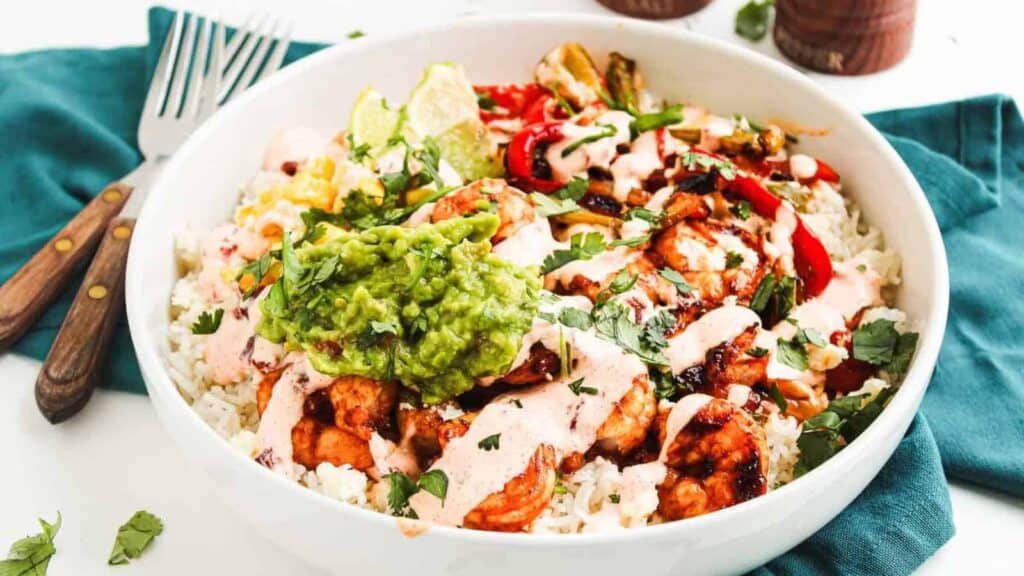 A bowl filled with rice, grilled chicken, sliced peppers, guacamole, corn, lime wedges, fresh cilantro, and a creamy sauce—perfect inspiration if you love seafood meals for everyday cooking—served with fork and knife on a teal napkin.