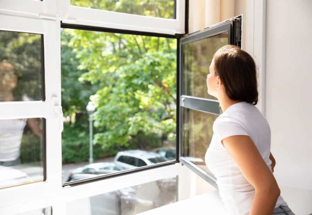 A woman in a white t-shirt looks outside through an open window; trees and parked cars are visible on a sunny day.