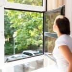 A woman in a white t-shirt looks outside through an open window; trees and parked cars are visible on a sunny day.