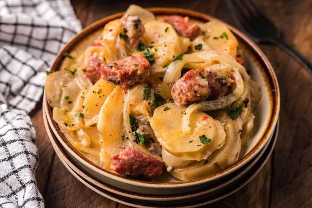 A bowl of creamy sliced potatoes baked with onions and sausage pieces, garnished with chopped parsley, on a wooden table next to a checkered napkin.