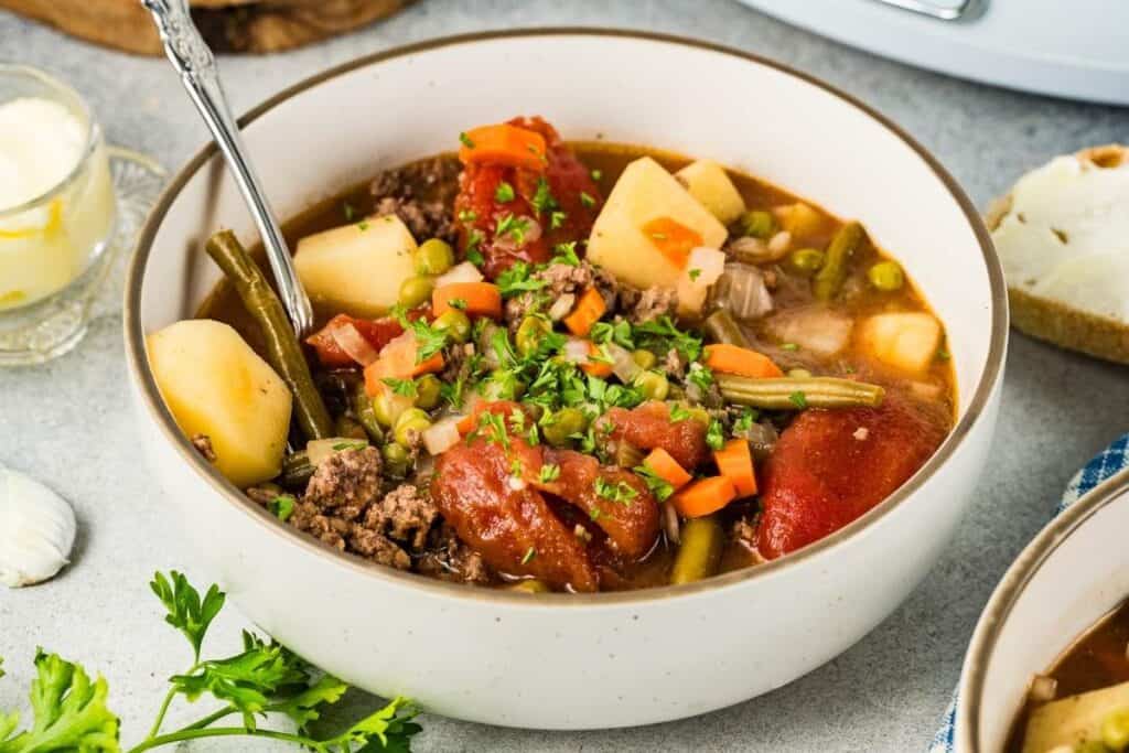 A bowl of beef and vegetable stew with potatoes, carrots, peas, green beans, and tomatoes, garnished with parsley, with a spoon in the bowl.