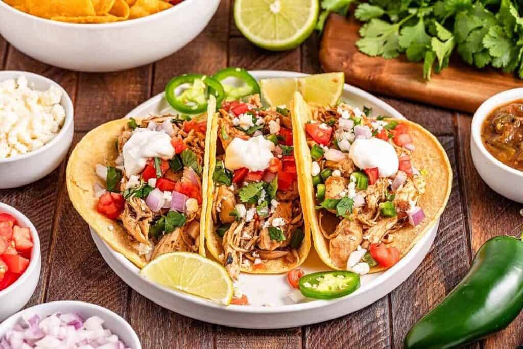 Three chicken tacos topped with chopped onions, tomatoes, cilantro, jalape&ntilde;os, and sour cream, served with lime wedges on a white plate, surrounded by side dishes.