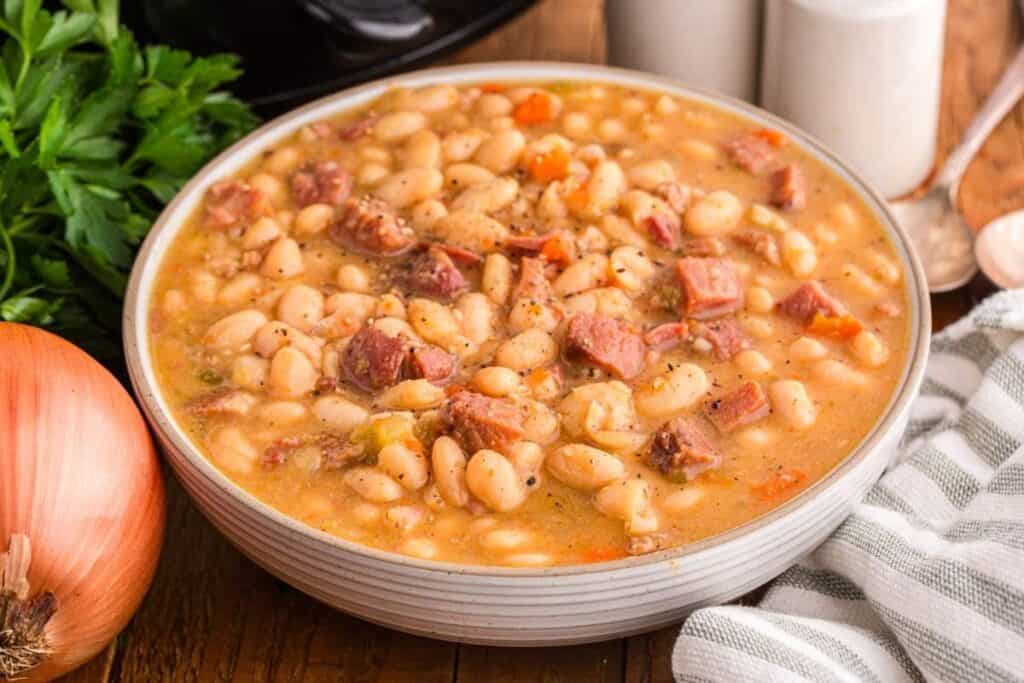 A bowl of white bean soup with chunks of ham and vegetables, perfect for fans of slow cooker soups and stews, sits on a wooden table next to an onion, parsley, and striped cloth.