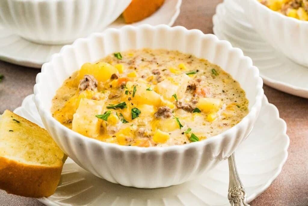A white bowl of creamy soup with ground meat, potatoes, corn, and herbs, served with a slice of bread on a white saucer and a spoon.