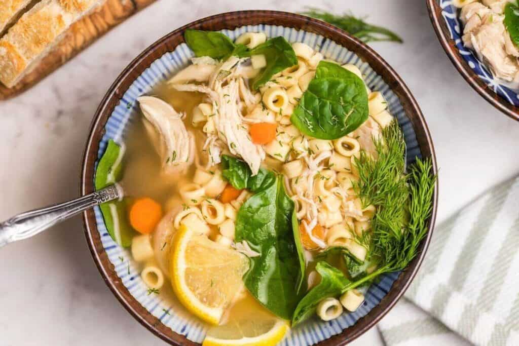A bowl of chicken noodle soup with shredded chicken, pasta, spinach, carrot slices, fresh dill, and lemon wedges, served with a spoon.