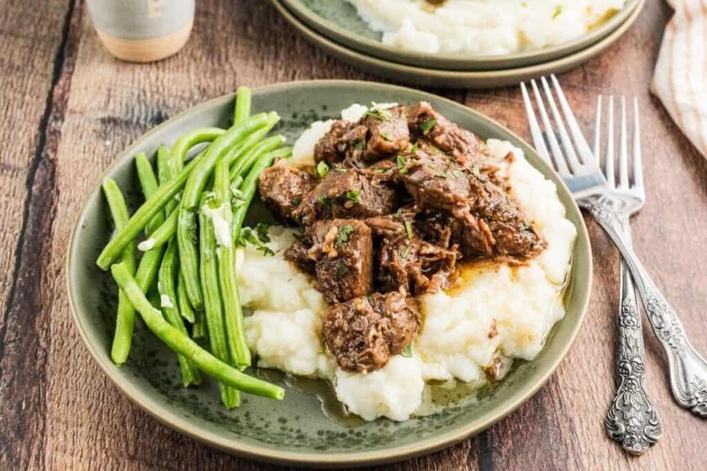 A plate of mashed potatoes topped with braised beef and a side of steamed green beans, inspired by tasty beef recipes, with forks beside the plate on a wooden table.