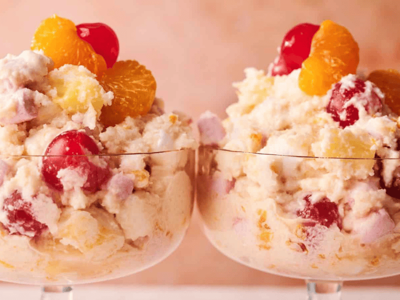 Two glass dessert cups filled with ambrosia salad, featuring whipped cream, cherries, pineapple, mandarin oranges, and marshmallows against a soft pink background.